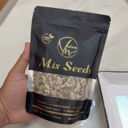 A hand holding a black stand-up pouch of Islam W Mix Seeds, showing the clear window with visible mixed seeds inside, set against a white marble countertop.